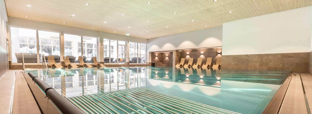 Innenansicht eines Schwimmbads mit Liegestühlen und großen Fenstern