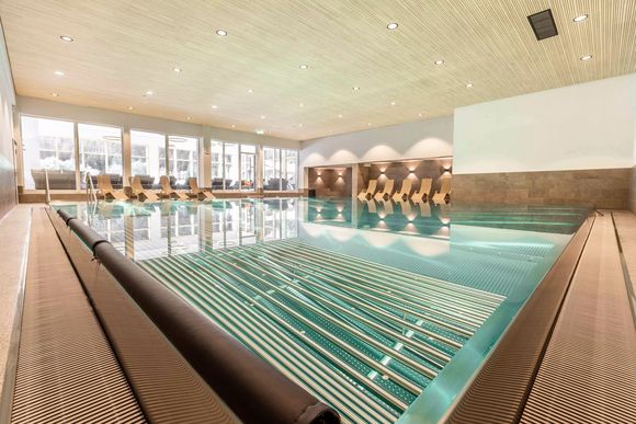 Innenansicht eines Schwimmbads mit Liegestühlen und großen Fenstern