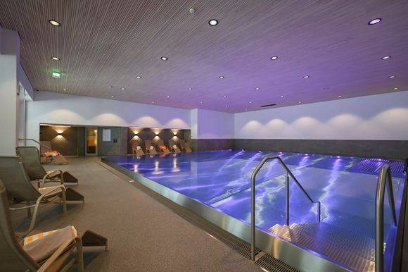 Innenpool mit blau beleuchtetem Wasser und Liegestühlen am Rand