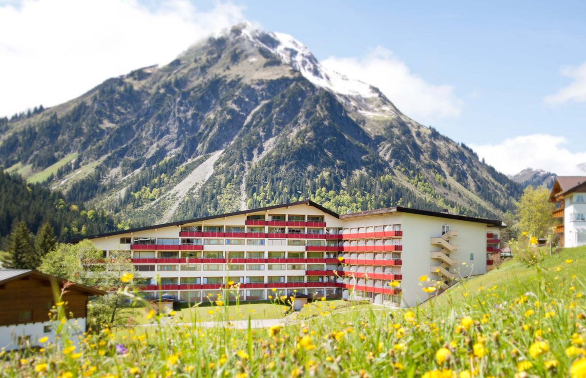 Hotelgebäude im Sommer mit roten Balkonbrüstungen vor einem Bergmassiv