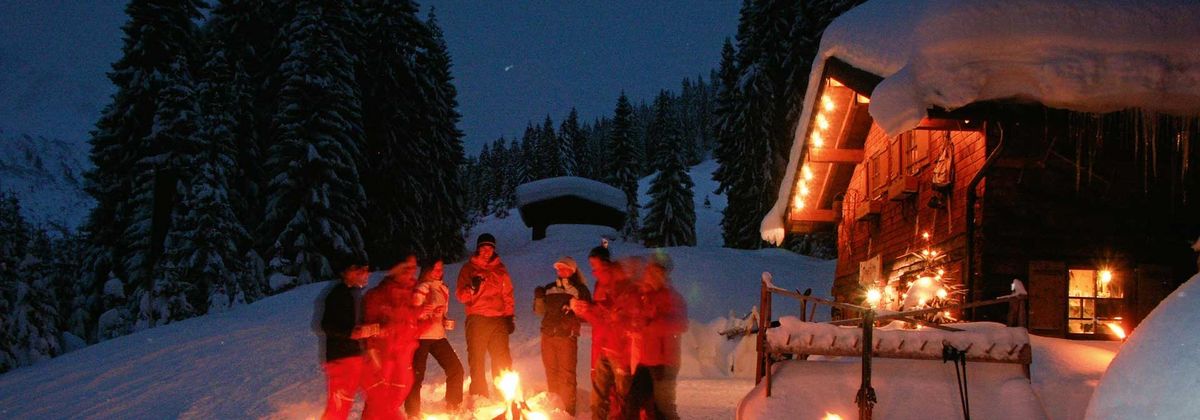 Gruppe von Menschen mit Fackeln vor einer verschneiten Hütte bei Nacht